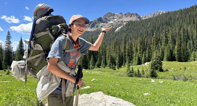 a student wearing a backpack stands in a green alpine meadow and pretends to hold the large mountain in the background in the palm of their hand. 